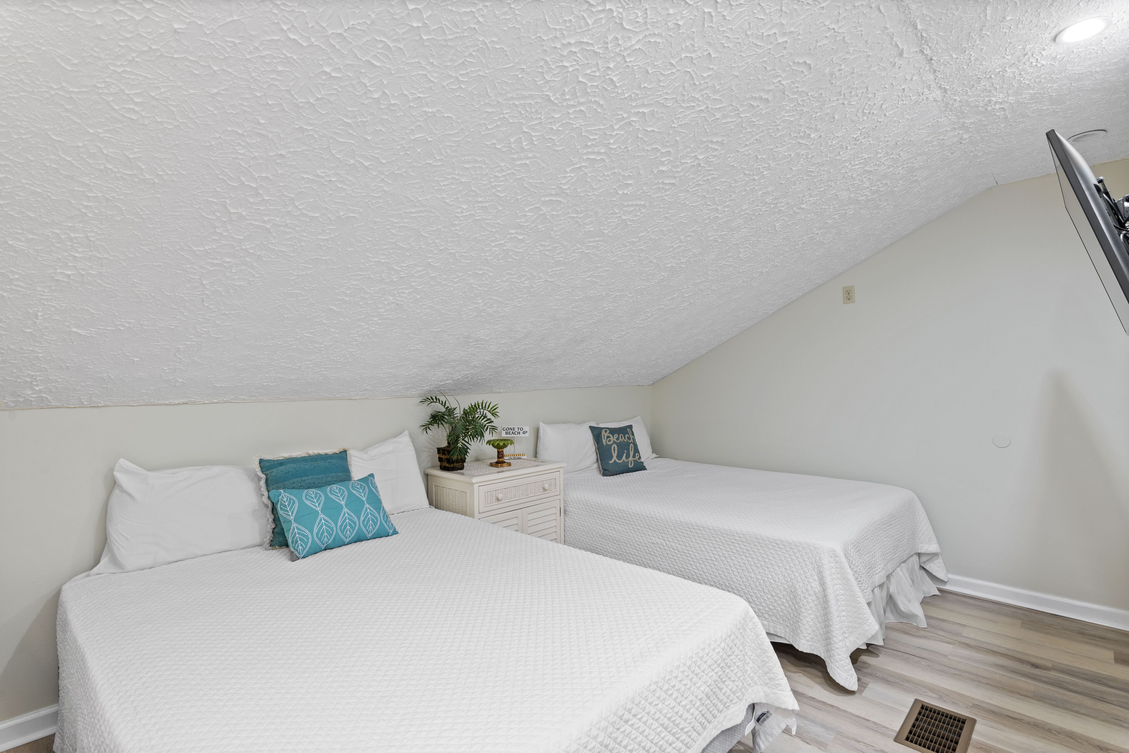 Une chambre avec deux lits, du linge de lit blanc et une petite commode agrémentée d’une plante.
