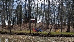 A forested area with a small cabin and colorful Adirondack chairs by a pond.
