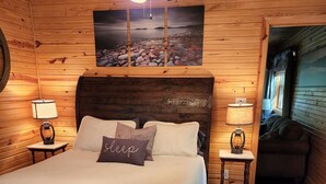 A wooden cabin bedroom with a large bed, two bedside lamps, a ceiling fan, and a view of the outside through a window.