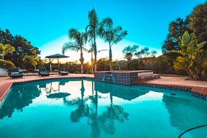Una piscina con borde de azulejos, rodeada de sillas de descanso y sombrillas, con palmeras y un cielo despejado al fondo.