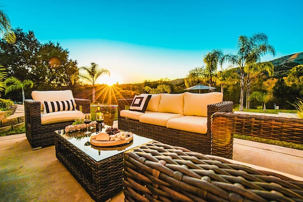 An outdoor seating area with wicker furniture and a glass-top coffee table, set against a backdrop of trees and a sunset.