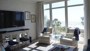 A living room with a large TV, comfortable seating, a coffee table, and a view of the ocean through large windows. - 3,000 Sq. Ft 2nd Floor 4 Bed/Bath End Unit  Ocean Side (Panama City Beach)