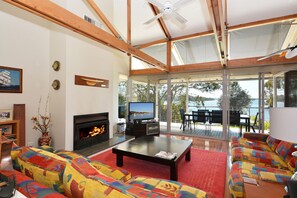 A spacious living room with a fireplace, a television, and a large window overlooking a body of water. - Edgewater Lakehouse at Morisset Memories (Morisset Park)
