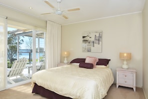 Une chambre à coucher comprenant un lit, deux tables de chevet avec des lampes, un ventilateur de plafond, une grande fenêtre avec des rideaux et une œuvre d’art accrochée au mur.