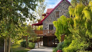 A stone house with a red roof and wooden deck, surrounded by lush greenery and trees.
