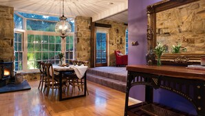 A cozy dining area with a stone fireplace, wooden dining table, and chairs. A large window offers a view of greenery outside.