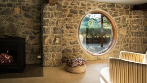 A stone-walled room with a fireplace, a round window with a view of greenery, and a woven pouf.