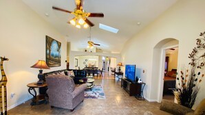 A spacious living area with a sofa, armchairs, a coffee table, and a television. There is a ceiling fan, a dining area in the background, and a decorative plant.