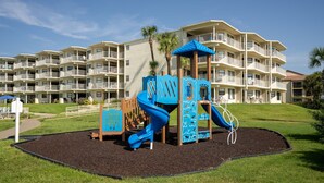 Ein Spielplatz mit einer blauen Rutsche und Kletterbereich, umgeben von einer angelegten Fläche mit Palmen.