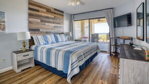 A bedroom with a bed, nightstand, wooden wall paneling, ceiling fan, TV, and a view of the outdoors. - CRC 3403 - Newly Renovated Direct Ocean View (St Augustine)