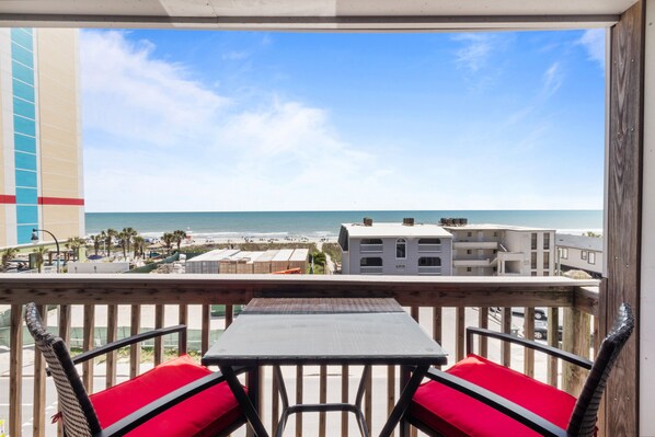 A balcony with a view of the beach and ocean, featuring a table and wicker chairs.