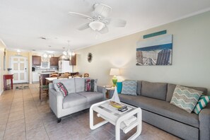 A modern living room with a grey sofa, a coffee table, and a ceiling fan. There is a kitchen in the background with wooden cabinets and a dining area.