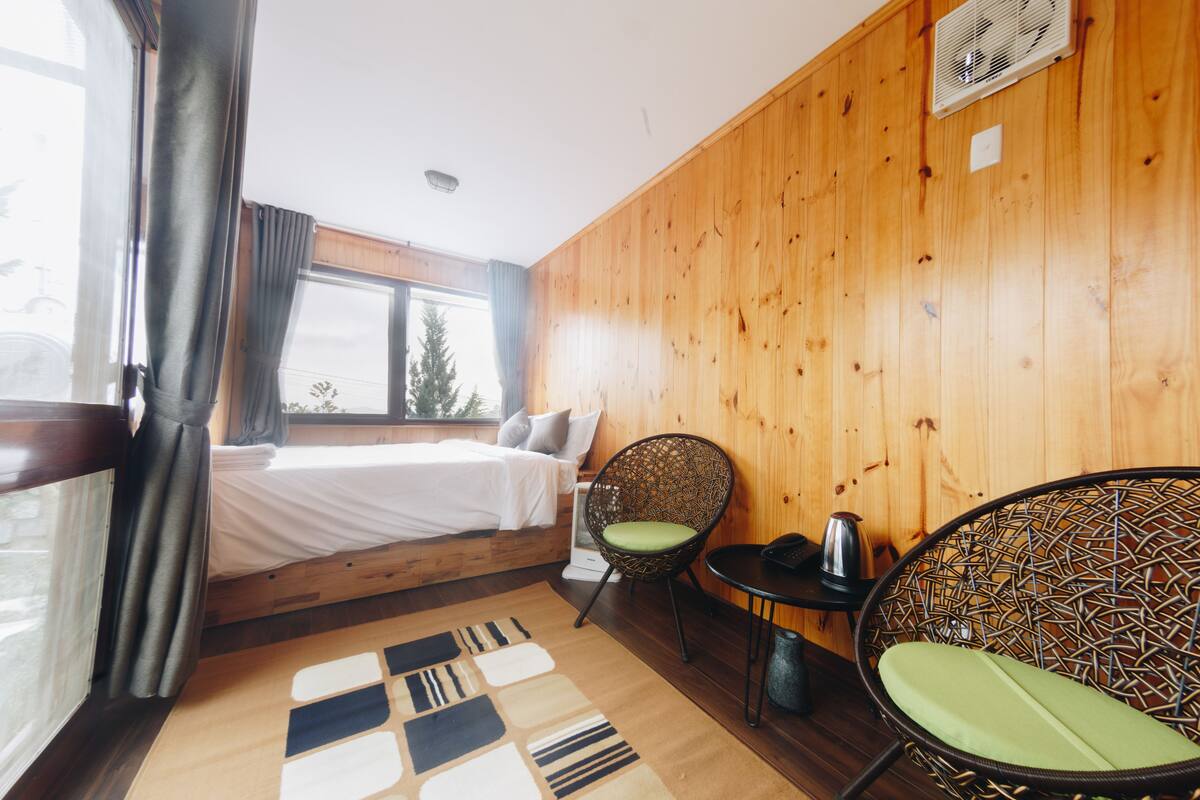 A wooden-paneled room with a bed, two wicker chairs, a small table, and a window with curtains.