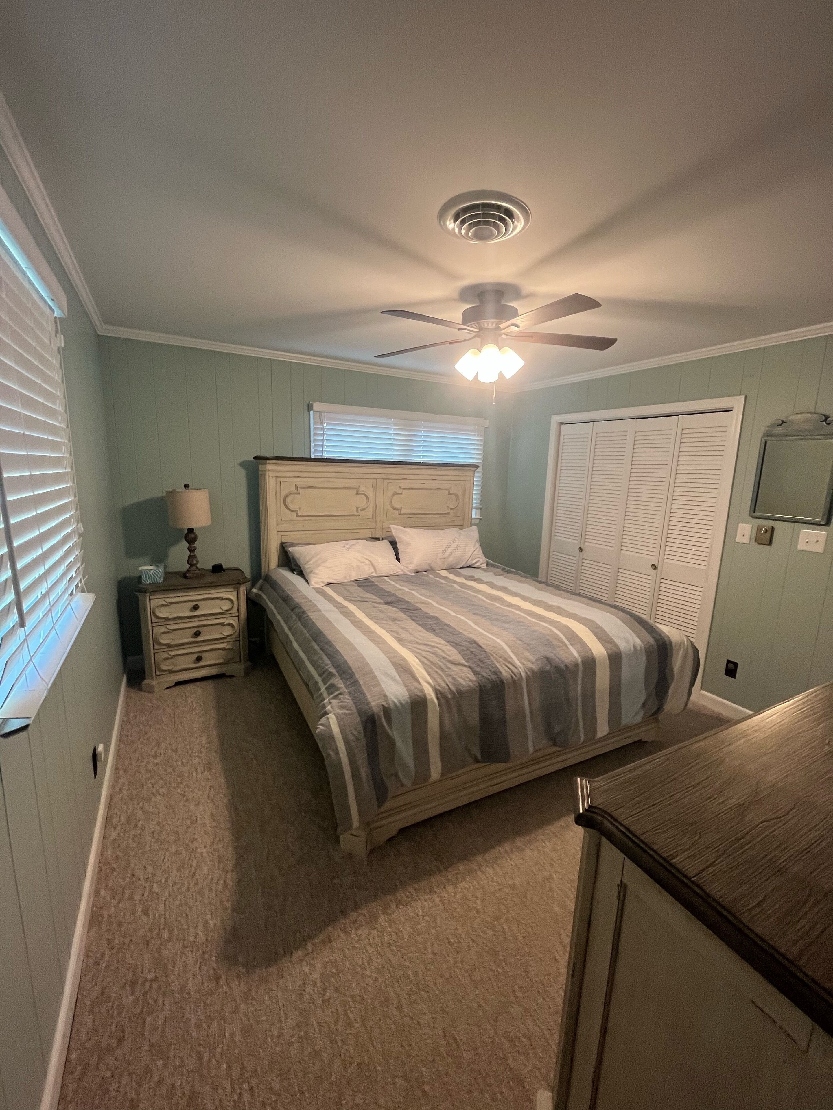 Une chambre à coucher comprenant un lit, une table de chevet, un ventilateur de plafond et une fenêtre avec des stores.