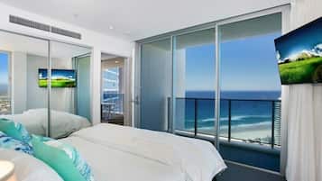 A modern hotel room with a large bed, a TV, a balcony with a sea view, and a sliding glass door.