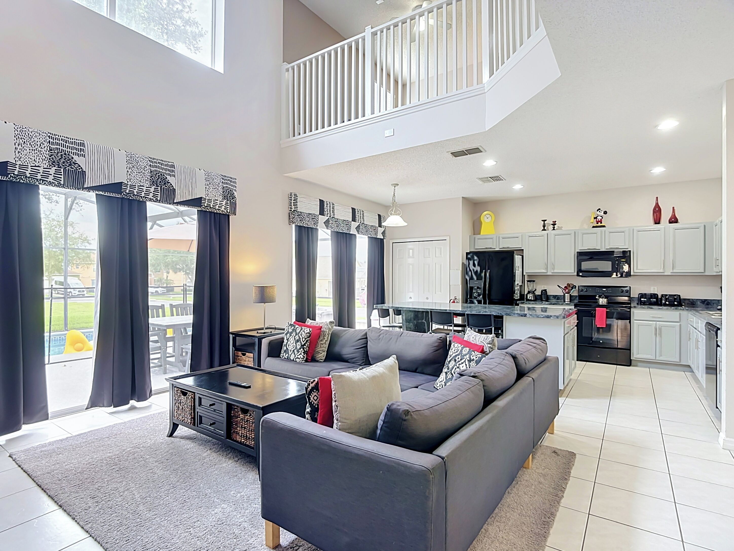 A modern living room with a sofa, coffee table, and curtains. A kitchen with appliances and cabinets is visible in the background.