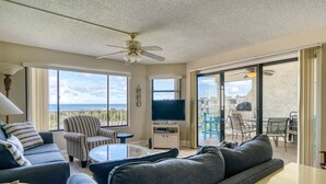 Una sala de estar moderna con sofá, sillón y televisor, ventanas amplias con vistas, y ventilador de techo.