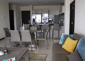 Un salon moderne avec une table à manger en verre, des sièges confortables, un canapé avec des coussins et un coin cuisine équipé d’appareils électroménagers.