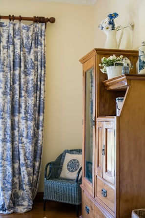 A room with a wooden cabinet, a patterned curtain, a chair with a cushion, and a vase with flowers.