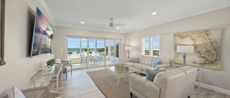 A spacious living room with a large sofa, a coffee table, and a dining area with chairs. There is a large window providing a view of the outdoors.