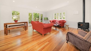 A spacious living room with hardwood flooring, a large window, a fireplace, and red upholstered furniture. - Poet's Cottage 1912 Clifftop Walks, Fire, Spa Bath (Wentworth Falls)
