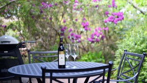 A round metal table with a bottle of wine and two glasses, surrounded by chairs, set outdoors with greenery and purple flowers in the background. - Poet's Cottage 1912 Clifftop Walks, Fire, Spa Bath (Wentworth Falls)