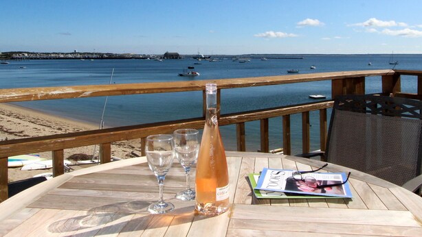 A wooden deck with a table featuring a bottle, glasses, and a magazine. The deck overlooks a beach and the sea with boats in the distance.
