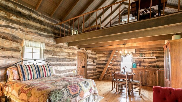 A rustic log cabin bedroom with a bed, a dining table, and a red armchair.