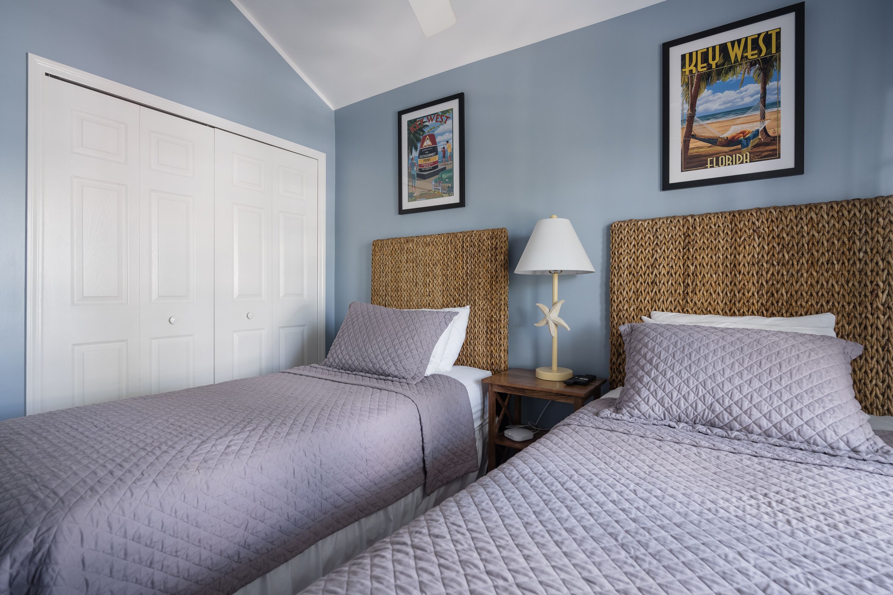 A bedroom with two beds, a white door, a framed picture of Key West, and a woven headboard.