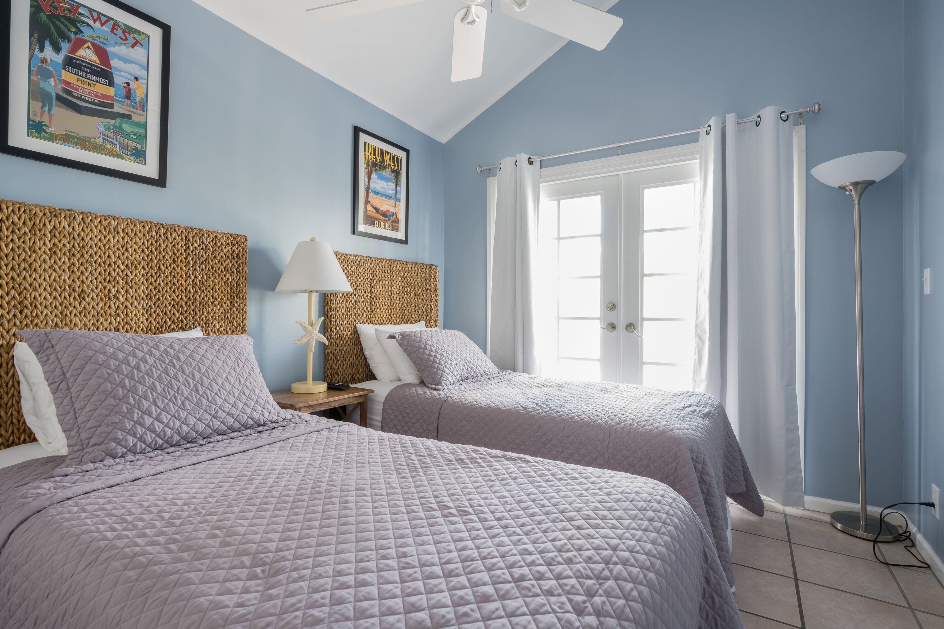 A room with two beds, a ceiling fan, a framed picture on the wall, and a floor lamp.