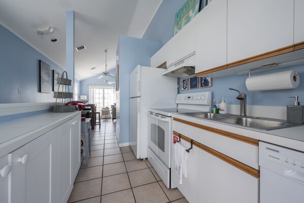 A modern kitchen with white cabinets, a built-in oven, and a dishwasher. There is a window with curtains, a ceiling fan, and a sink with a soap dispenser.