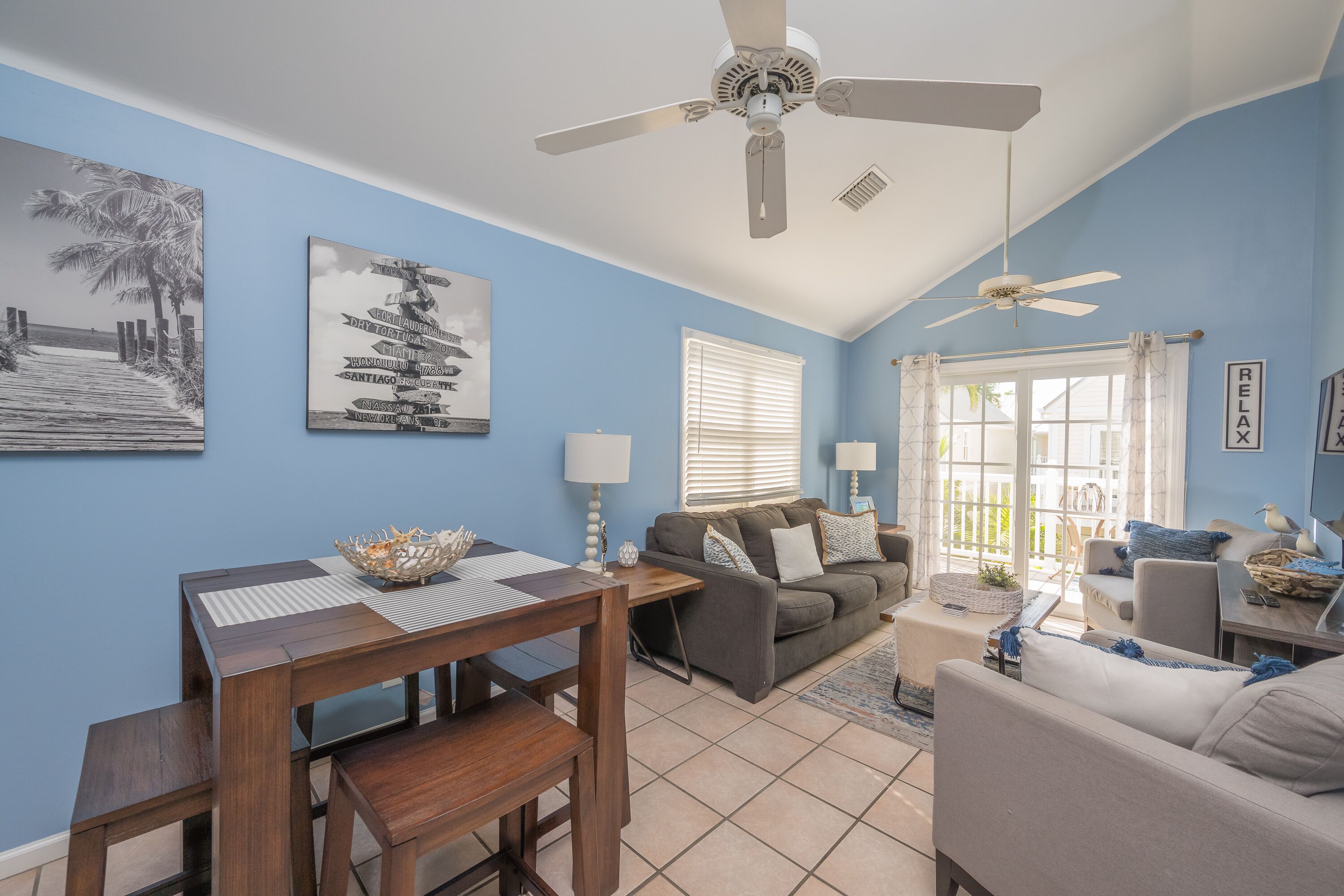A living room with a dining table, sofa, and ceiling fan. There are wall art pieces and a window with blinds.