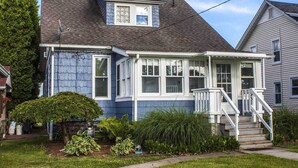 Une maison à deux étages avec un porche à l’avant, un petit jardin et une cheminée bien visible.