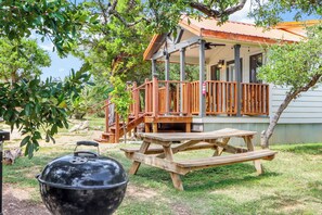Una terraza de madera con una mesa de picnic y una parrilla, rodeada de árboles y una casa con porche.