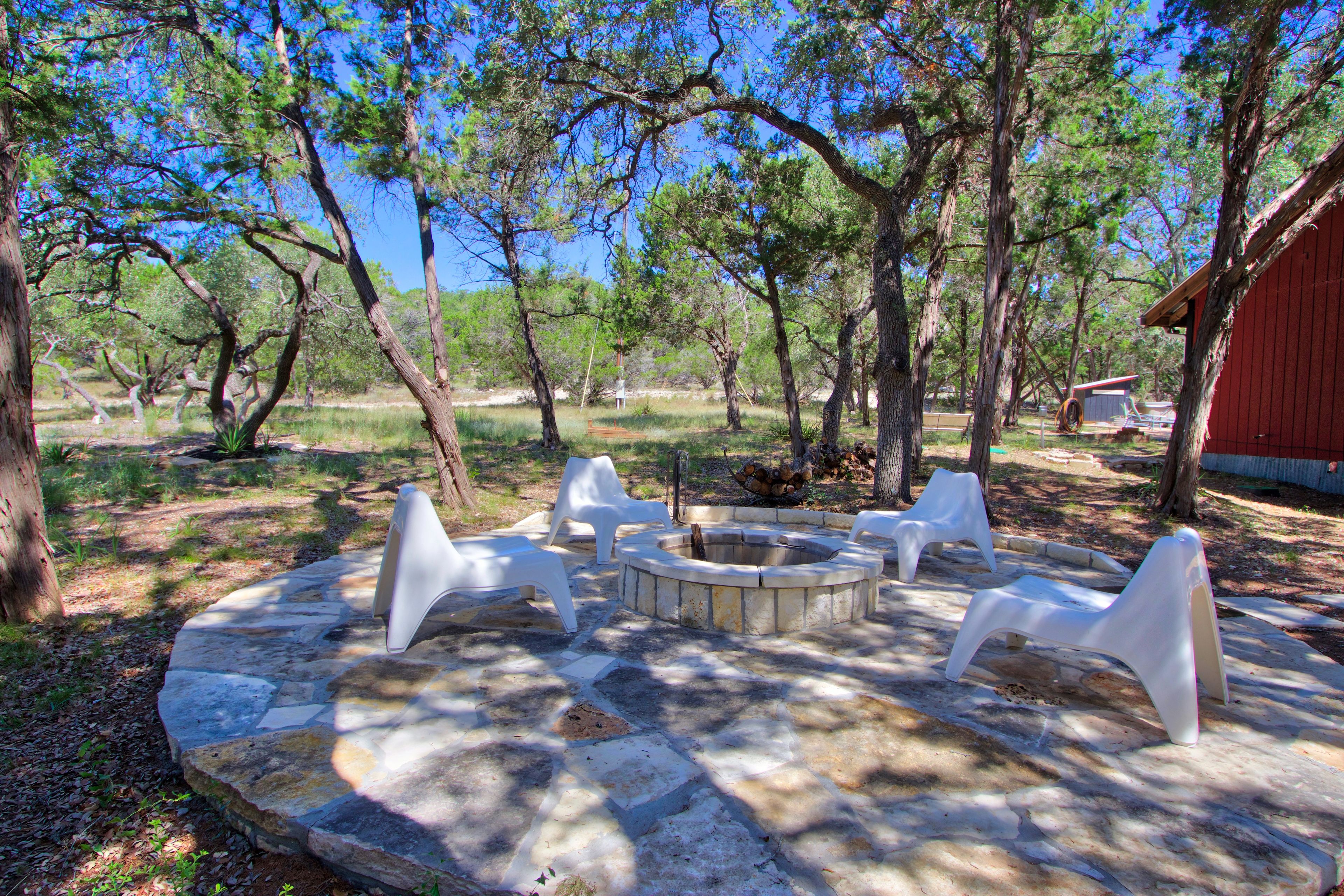 A stone fire pit with white plastic chairs, surrounded by trees in a wooded area.