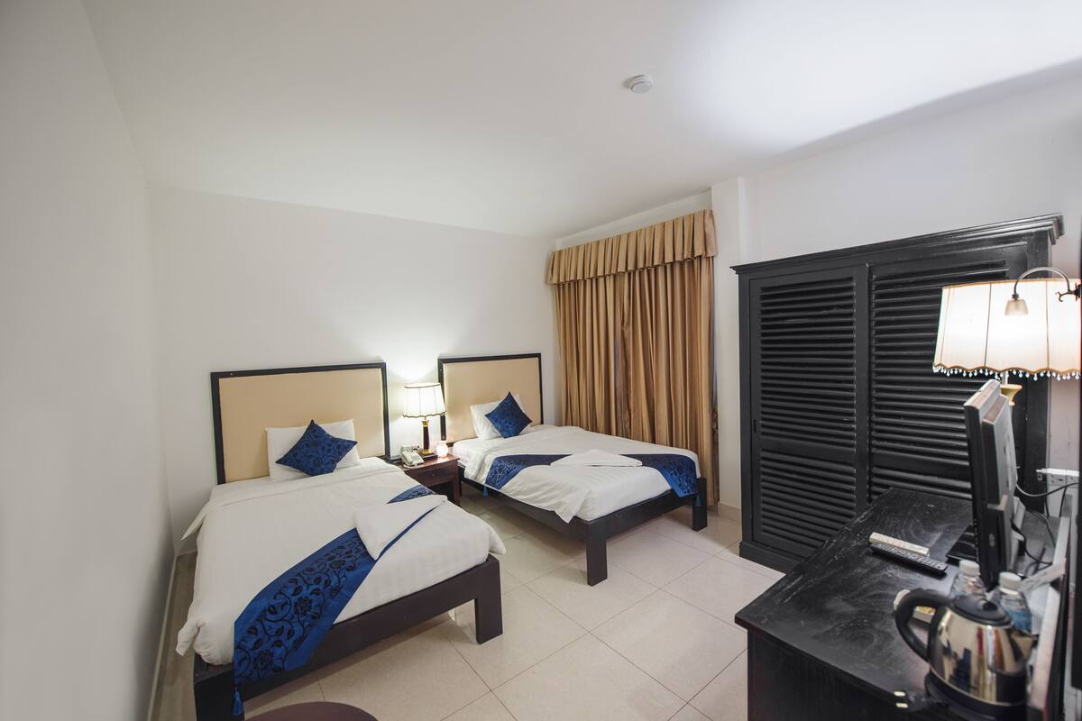 Habitación de hotel con dos camas, un escritorio con computadora, una tetera y una ventana con cortinas.