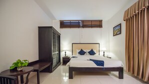 Une chambre d’hôtel comprenant un lit, des tables de chevet, un bureau, une chaise, une plante en pot et une fenêtre avec des stores.