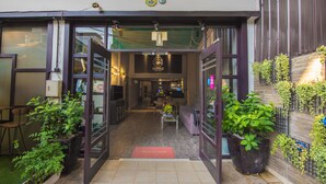 L’entrée d’un café, avec un paillasson accueillant, des plantes en pot et un intérieur visible, comprenant des tables et un comptoir.