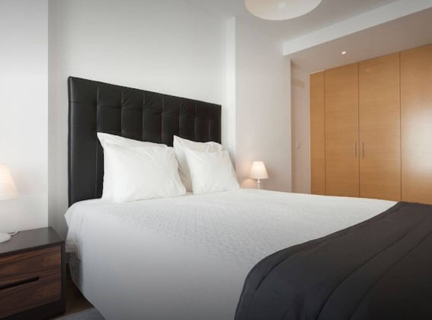 A modern bedroom with a large bed, dark headboard, white bedding, and a wooden wardrobe.