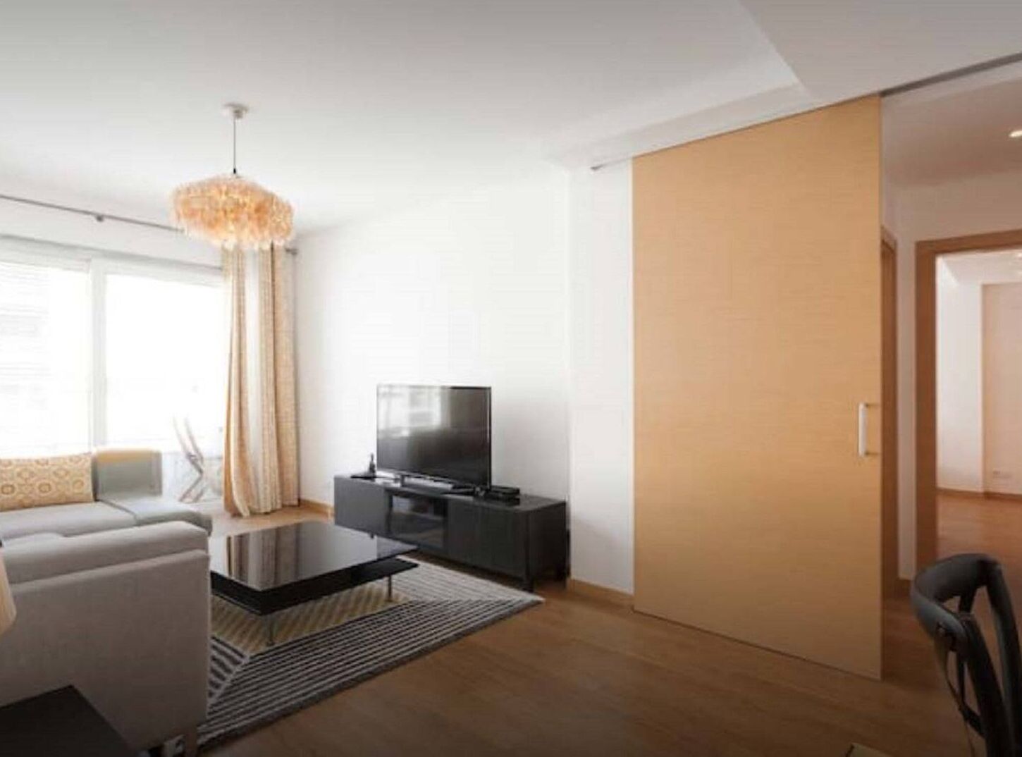 Uma sala de estar moderna com televisão de tela plana, uma mesa de centro de vidro, um sofá e uma janela grande com cortinas.