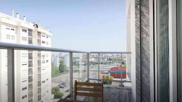 A balcony with a wooden bench and a small table, overlooking a cityscape with buildings and a road.