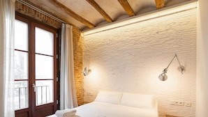 A hotel room with a large bed, white linens, a wooden bench, and a brick wall.