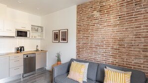 Eine moderne Küche mit weißen Schränken, einer Mikrowelle und einem Geschirrspüler. Ein graues Sofa mit gestreiften Kissen lehnt an einer Ziegelwand.