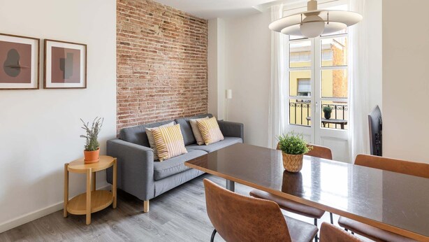 Ein modernes Wohnzimmer mit einer Ziegelwand, einem grauen Sofa, einem Holzesstisch und einem großen Fenster mit Blick auf Gebäude.
