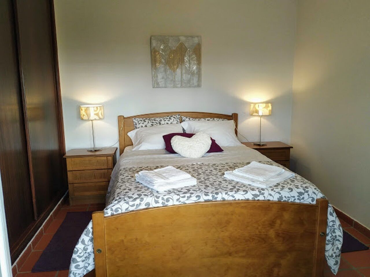 Un lit bien fait, avec une couvre-lit à motifs, deux lampes de chevet et un coussin en forme de cœur.