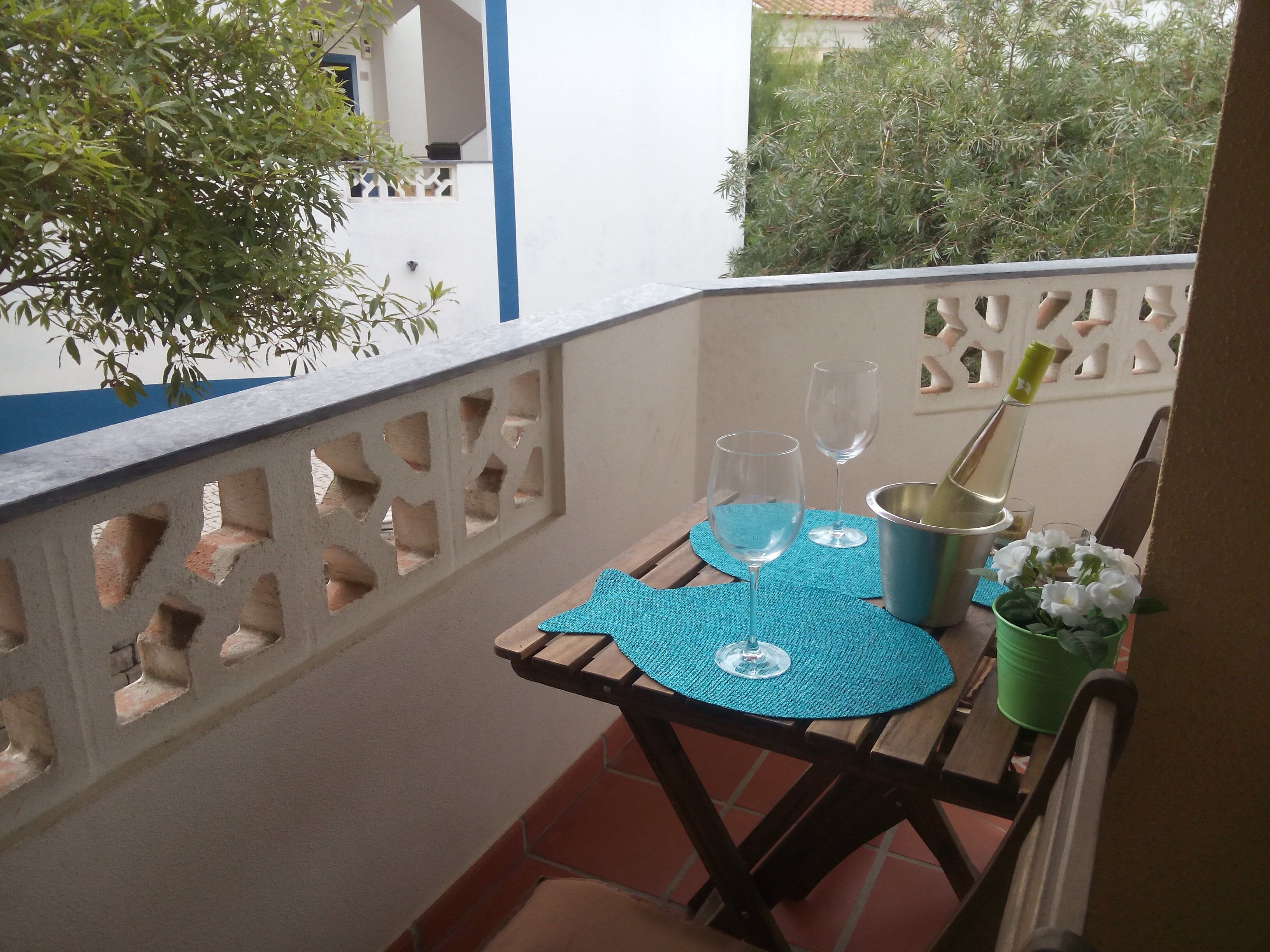 Uma varanda com mesa posta para dois, com dois copos de vinho, uma garrafa e um vaso verde com flores.