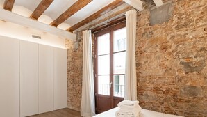 A room with a wooden ceiling, stone walls, a large window with curtains, a bed with white linens, and a wardrobe.