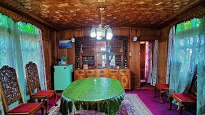 A room with a round table covered with a green tablecloth, surrounded by wooden chairs with red cushions. The room features a patterned carpet, wooden walls, and a ceiling with a geometric design. - Shiraz Deluxe Houseboat (Srinagar)