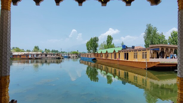 A view through an ornate window with geometric patterns, overlooking a calm river with boats and traditional buildings. - Shiraz Deluxe Houseboat (Srinagar)