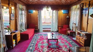 A room with a patterned ceiling, a red carpet, a wooden desk, a red sofa, and a large window with curtains. - Shiraz Deluxe Houseboat (Srinagar)